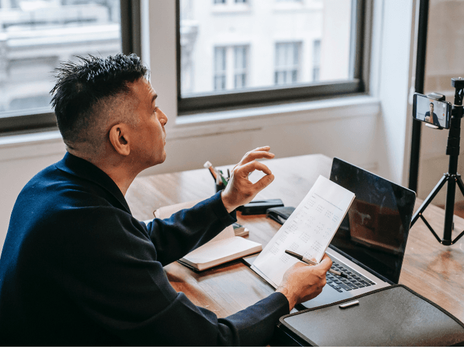 Man giving virtual presentation in front of computer and phone holding notes