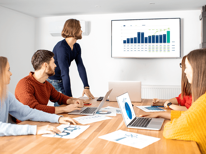 person presenting bar chart of data on tv to group of people seated around a large table