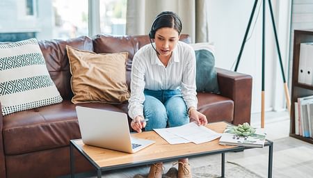 woman sitting on leather couch wearing headset while typing on laptop and looking at papers.