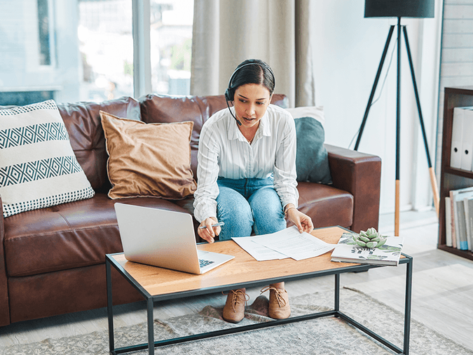 woman sitting on leather couch wearing headset while typing on laptop and looking at papers.