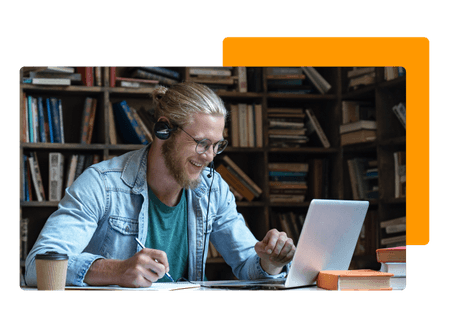 man sitting at desk wearing headset laughing while writing notes and looking at laptop with full bookshelf in background