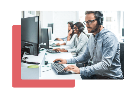 man wearing headset in a call centre sitting at his desk with two women in the background wearing headsets sitting at their desks