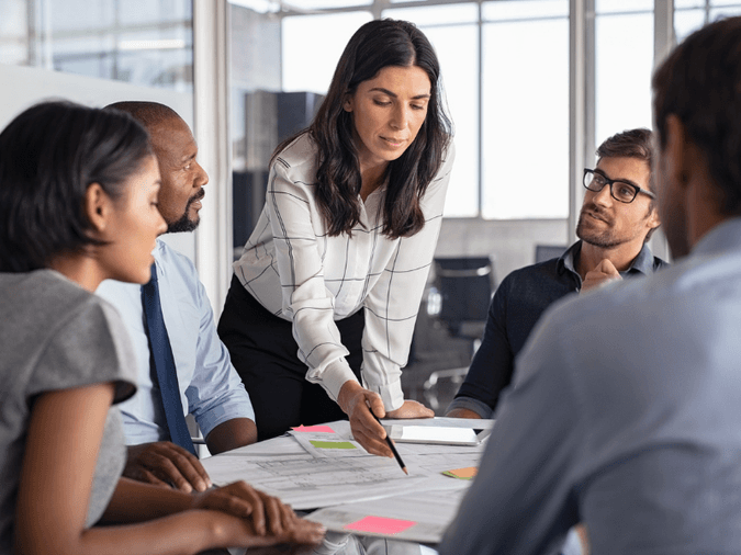 woman presenting to business people around a table and pointing a pen towards text on a stack of documents