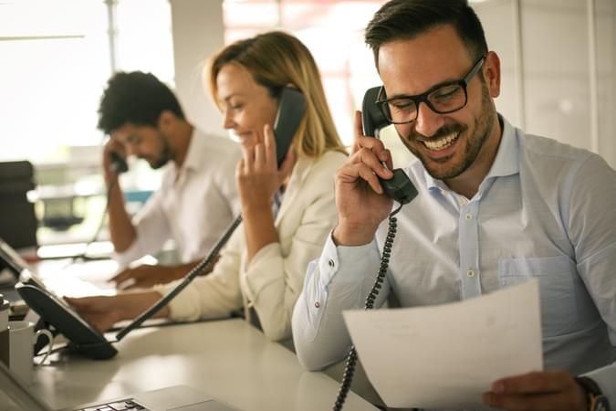 A man on a desk phone handset with his two colleagues in the background also on desk handsets - net2phone Canada - Business VoIP Phone System