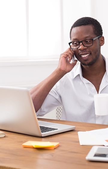 A man at his desk on the phone in front of his laptop at home