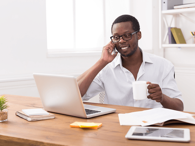 A man at his desk on the phone in front of his laptop at home