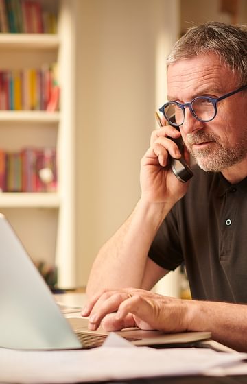 man typing on laptop while speaking on cordless phone seated at desk in front of a book case