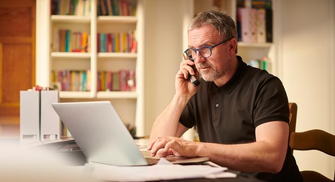 man typing on laptop while speaking on cordless phone seated at desk in front of a book case