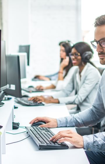 man on headset sitting at his desk top in a call centre with two women in the background wearing headsets sitting at their desks