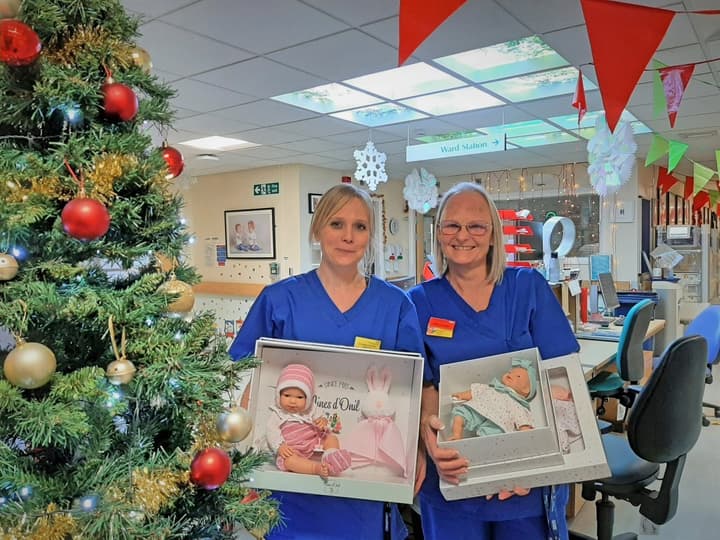 Two female nurses holding boxes of dolls next to a Christmas tree in a hospital ward.