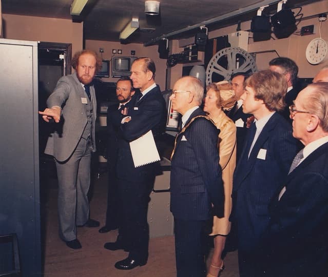 A man pointing to an equipment rack in front of a group of 10 people in formal dressing
