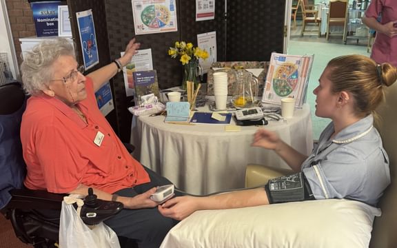 Care home resident talks to carer about information on a display board whilst taking her blood pressure