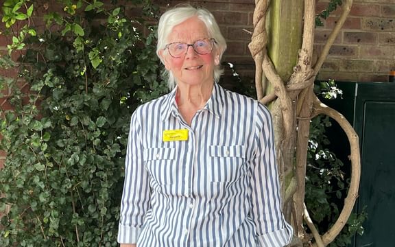Mid shot of resident Marjorie standing in front of a gnarled tree trunk in the Cotman House garden