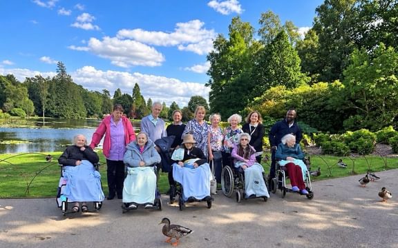 Group of residents in wheelchairs with carers outside in front of lake and trees