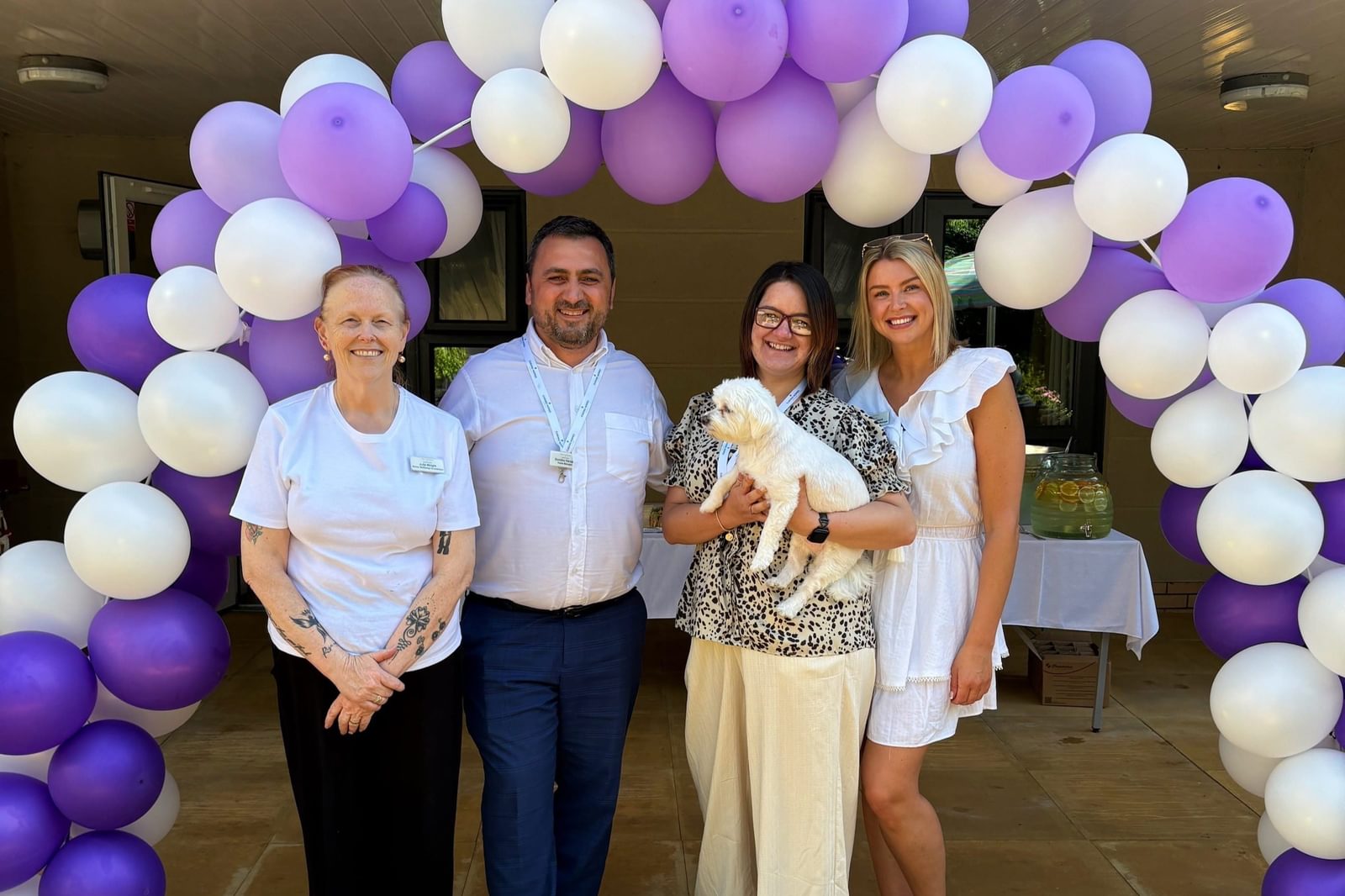 Care Home team and white dog pose under a purple balloon arch