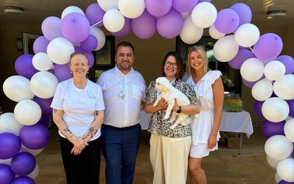 Care Home team and white dog pose under a purple balloon arch