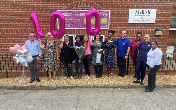 Three women standing next to each other holding pink balloons that spell out 100.