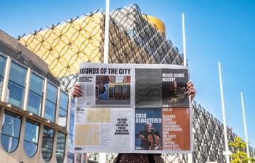 Photograph of somebody reading the CBSO newspaper outside Birmingham Central Library and the Birmingham Repertory Theatre
