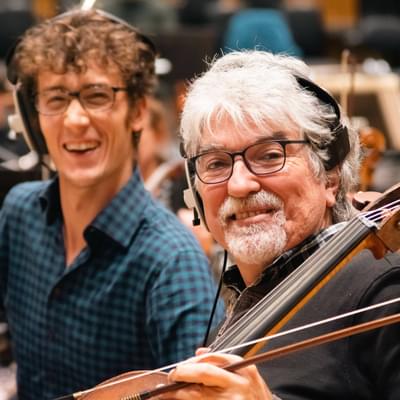 Photograph of cellist Eduardo Vassallo smiling at the camera during a recording session whilst he wears headphones and holds his cello