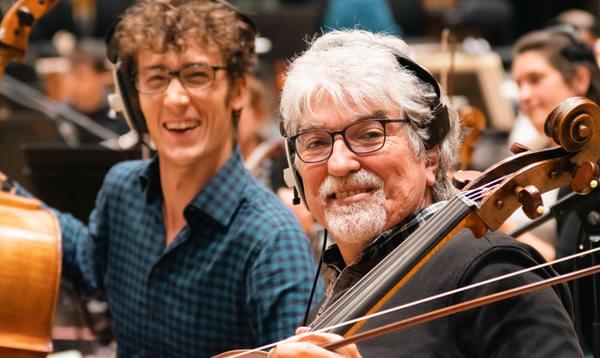 Photograph of cellist Eduardo Vassallo smiling at the camera during a recording session whilst he wears headphones and holds his cello