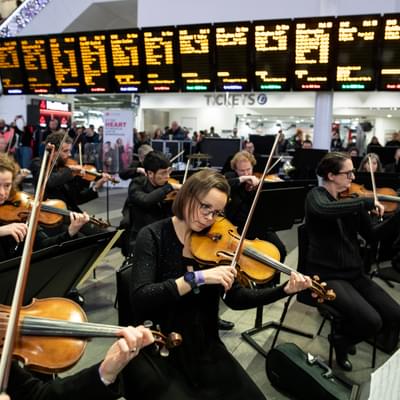 Photograph of the orchestra performing beneath the departure boards at New Street Station