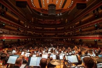 The Orchestra play on-stage at Symphony Hall. Behind the conductor, the hall is full of audience.