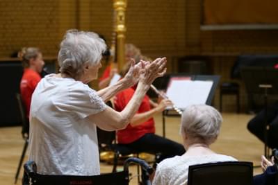 Photograph of an elderly woman giving a standing ovation to a small ensemble of players