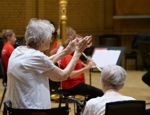 Photograph of an elderly woman giving a standing ovation to a small ensemble of players