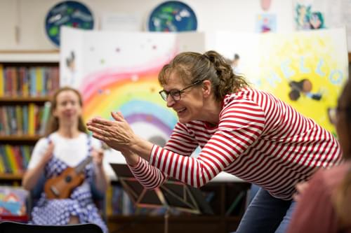 Photograph of Kate Setterfield clapping and smiling in a children's library, whilst another performer plays the Ukulele behind her
