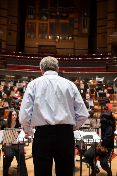 A back view of Mark Goodchild conducting an orchestra of young people on the Symphony Hall stage. Mark is wearing black trousers and a white shirt.