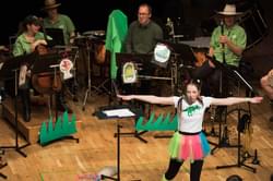 Bryony Morrison is standing on the CBSO Centre stage with a white t-shirt and a brightly coloured skirt. Her arms are stretched out from side to side. Behind her is a group of musicians dressed in green and brown safari costumes.