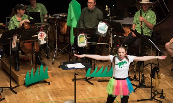Bryony Morrison is standing on the CBSO Centre stage with a white t-shirt and a brightly coloured skirt. Her arms are stretched out from side to side. Behind her is a group of musicians dressed in green and brown safari costumes.