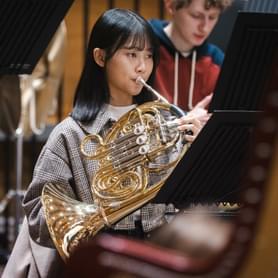Photograph of a young French horn player. She is playing the French horn during a rehearsal.