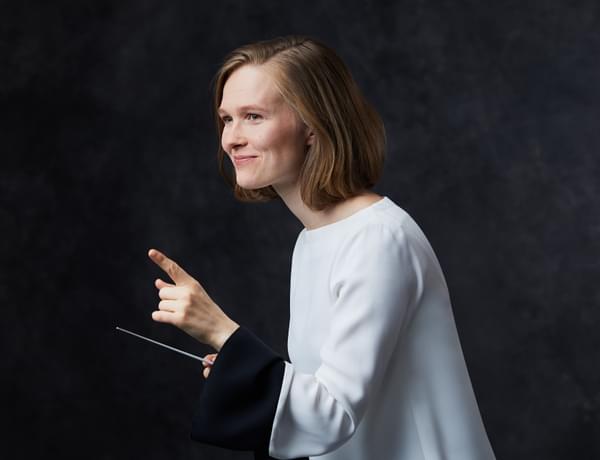 Conductor Mirga Gražinytė-Tyla stands in front of a dark background. She holds a white baton in her right hand and points with her left hand. She is smiling. She has medium length light brown hair, and is wearing a shite shirt.