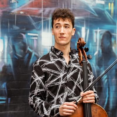 Photograph of Arthur Boutillier holding his cello in front of a wall decorated with graffiti