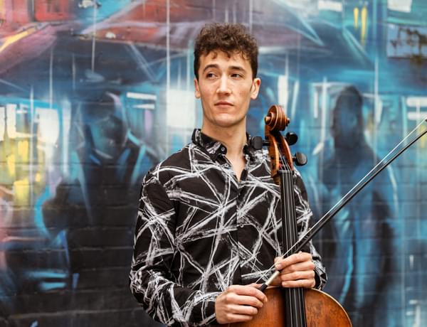 Photograph of Arthur Boutillier holding his cello in front of a wall decorated with graffiti