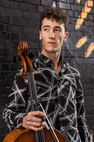 Photograph of cellist Arthur Boutillier. Arthur is leaning against a graffitied wall holding his cello.