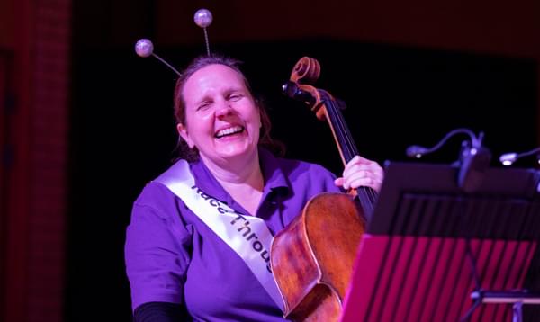 Photograph of cellist Helen Edgar laughing whilst performing in a Key Stage 1 Schools' Concert. She is wearing a sash and a headband with baubles on it.
