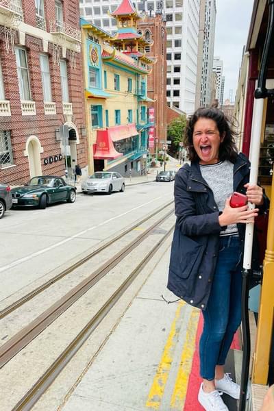 Jess Tickle is stood on the side of an open tram. She is windswept and smiling excitedly. There are city buildings in the background of the image.