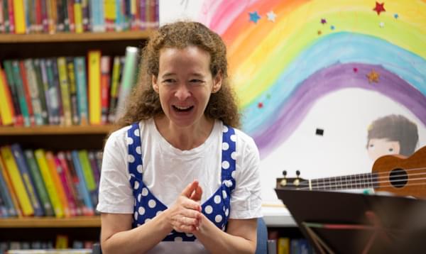 Photograph of violinist Julia Åberg laughing and clapping in front of a bookcase and an illustration of a rainbow