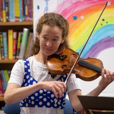 Photograph of Julia Åberg playing the violin in front of a bookcase and an illustration of a rainbow