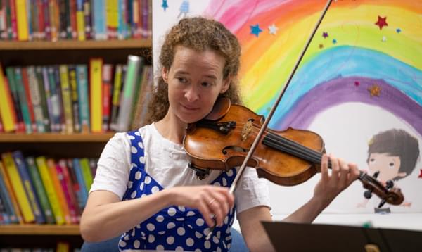 Photograph of Julia Åberg playing the violin in front of a bookcase and an illustration of a rainbow