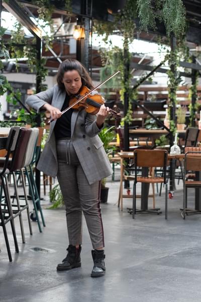 Colette Overdijk is playing her violin in Hockley Social Club. She is surrounded by chairs and tables, and greenery.