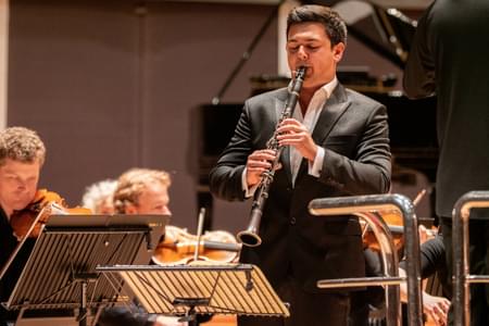 Oliver Janes is playing the clarinet during a concert. He is stood at the front of the orchestra.