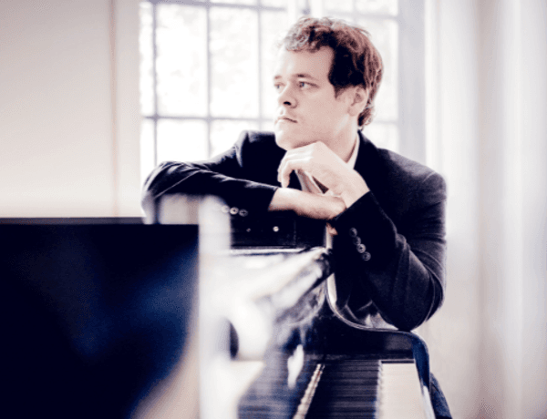 Photo of Benjamin Grosvenor sat beside the piano. Benjamin is leaning against the piano and looking away from the camera.