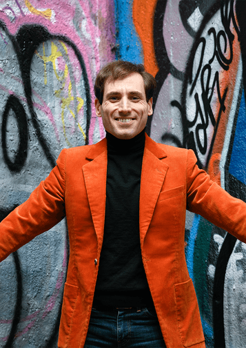 Photograph of Boris Giltburg posing in front of graffiti whilst wearing a bright orange jacket.
