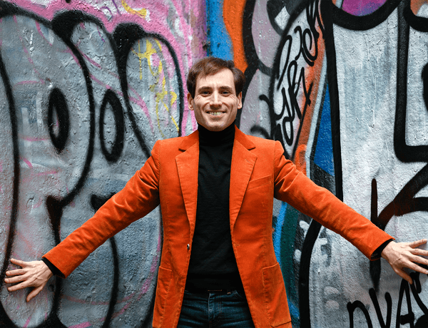 Photograph of Boris Giltburg posing in front of graffiti whilst wearing a bright orange jacket.