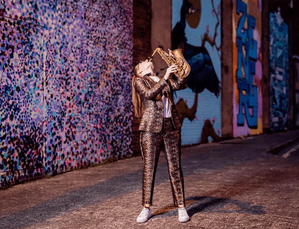 Photograph of Jess Gillam wearing a brightly patterned suit and playing the saxophone in front of graffiti.
