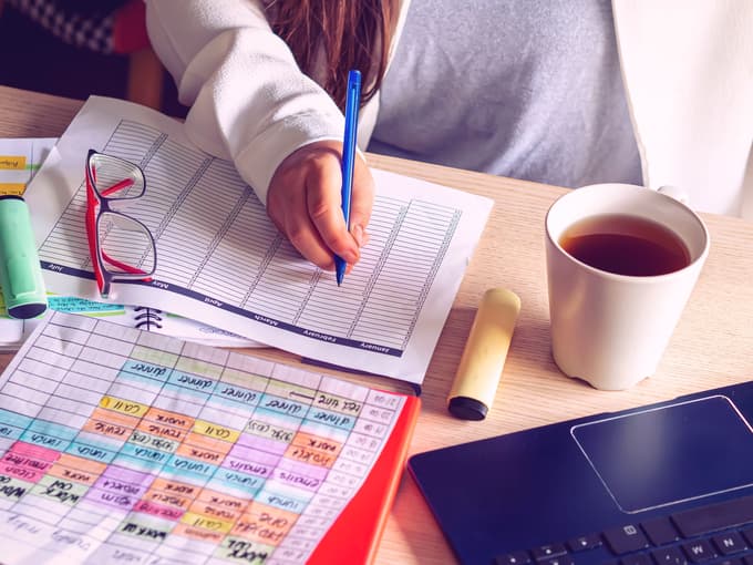 A person sitting at a table full of printed spreadsheets and various pens, glasses, a cup of coffee, and a laptop.