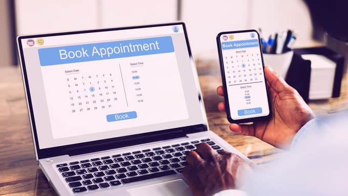 A behind-the-shoulders view of someone looking at a laptop and a phone with a "Book Appointment" webpage on the screens.